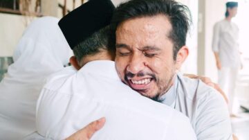 رجلان يحتضنان بعضهما بابتسامة، يرتدي أحدهما عمامة سوداء والآخر ملابس بيضاء. يظهر تعبير الفرح والتواصل العاطفي في المشهد، مع وجود شخص ثالث في الخلفية. تعكس الصورة لحظة مؤثرة من الألفة والمحبة.