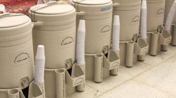 أكواب بلاستيكية موضوعة بجانب حاويات مياه كبيرة بلون بيج، في مكان يتسم بالنظافة والترتيب، مع سجادة حمراء في الخلفية.