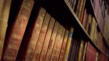 رفوف مكتبة تحتوي على مجموعة من الكتب القديمة ذات الغلاف الجلدي الملون، مع تفاصيل ذهبية على الأكتاف، تظهر في تركيز ضبابي.