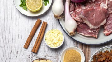 Ingredients for a savory dish arranged on a wooden surface, including raw meat, shallots, lemon slices, cinnamon sticks, butter, ground spices, and fresh herbs.