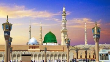 مسجد النبي في المدينة المنورة، يظهر القبة الخضراء والمآذن الشاهقة تحت سماء ملونة، مع وجود زوار يتجولون في الساحة.