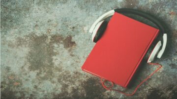 كتاب أحمر مع سماعات رأس بيضاء على سطح ملون، يرمز إلى تجربة الاستماع للكتب الصوتية.