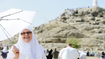 امرأة مسنّة ترتدي حجابًا أبيض وهي تحمل مظلة بيضاء، مبتسمة أمام جبل عرفة خلال موسم الحج. يظهر في الخلفية مجموعة من الحجاج. تعكس الصورة روح الفرح والتقوى في مناسك الحج.