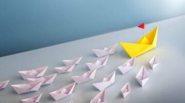 قارب ورقي أصفر يحمل علمًا أحمر يتقدم في مجموعة من قوارب ورقية بيضاء صغيرة على سطح أملس. الخلفية زرقاء فاتحة، مما يبرز القوارب الورقية بتصميم فني مبتكر.