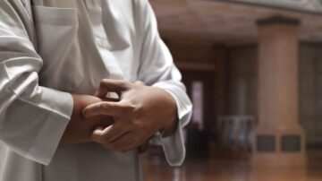 رجل يرتدي ثوباً تقليدياً، يقف في مكان داخلي، مع التركيز على يديه وهو يقوم بحركة تشير إلى الاستعداد أو التفكير. الخلفية تحتوي على عناصر معمارية تقليدية.