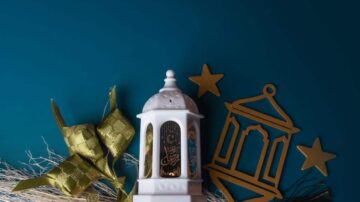 فانوس أبيض مزخرف بجوار قطع ذهبية على خلفية زرقاء، مع زخارف تقليدية تعكس أجواء الاحتفال.