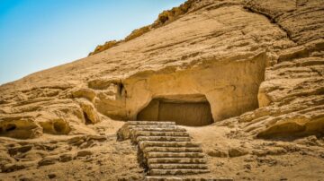 مدخل كهف محفور في جرف رملي مع درجات حجرية تؤدي إليه، تحيط به تضاريس صحراوية.