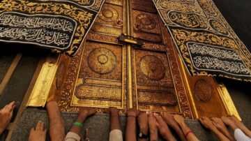 أبواب الكعبة المشرفة المزخرفة بالذهب، مع أيدٍ متعددة تلمس الأبواب. تظهر الكتابات العربية المنقوشة على الأبواب، مما يعكس الروحانية والتقدير في الموقع المقدس.