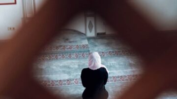 امرأة ترتدي الحجاب تصلي في مسجد، تظهر من خلال إطار نافذة. تصميم السجادة يظهر تفاصيل زخرفية. الإضاءة الطبيعية تخلق جوًا هادئًا.