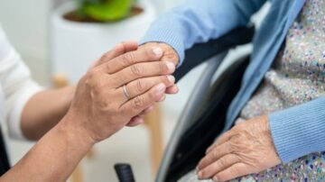 يد مريضة مسنّة تمسك بيد مقدّم الرعاية في بيئة دافئة. تعكس الصورة لحظة حميمية ودعماً عاطفياً، مما يبرز أهمية الرعاية الصحية للمسنين.