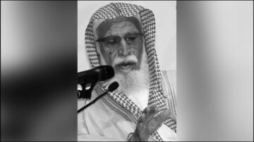 رجل مسن يرتدي لباس تقليدي سعودي، يتحدث في ميكروفون، يظهر تعبير جاد. شعره أبيض ولحيته طويلة، ويعتمر غطاء رأس مزخرف. الخلفية بسيطة وغير ملونة.