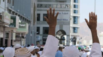 أشخاص يرتدون ملابس تقليدية إسلامية يرفعون أيديهم في شارع مزدحم، مع مباني حديثة في الخلفية. تعكس الصورة أجواء الحج أو العمرة في مكة، حيث يتجمع الحجاج في المكان.