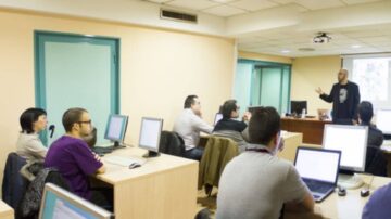 فصل دراسي مع مجموعة من الطلاب يجلسون أمام أجهزة الكمبيوتر بينما يقوم المعلم بإلقاء محاضرة. يركز الطلاب على الشرح، مع وجود لوحات بيضاء في الخلفية.