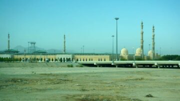 منظر لمدينة مكة المكرمة يظهر فيها المسجد الحرام والجبال المحيطة، مع عمارات حديثة ومآذن بارزة. السماء زرقاء وصافية، وتظهر الأرض القاحلة في المقدمة.