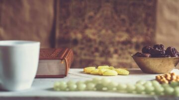 مائدة إفطار تتضمن فنجان قهوة أبيض، وكتاب، ووعاء نحاسي يحتوي على تمر، وحلويات ملونة، مع خلفية منسوجة تقليدية. تعكس الصورة أجواء رمضانية مميزة.