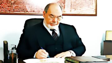 رجل مسن يرتدي بدلة رسمية يجلس على مكتب، يكتب بمساعدة قلم وأوراق. الخلفية تعرض جداراً مزخرفاً.