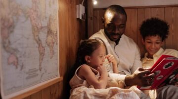 رجل يقرأ كتابًا لأطفال في زاوية دافئة، مع خريطة العالم معلقة على الجدار. الأطفال يظهرون مستمتعين بالقراءة، مع تعابير وجه سعيدة.