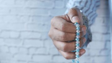 يد ممدودة تحمل مسبحة زجاجية بألوان زرقاء وشفافة، مع خلفية من طوب أبيض.