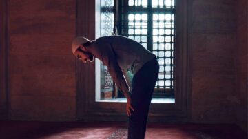 رجل مسلم يؤدي الصلاة في مسجد، يركع أمام نافذة مضاءة، مع التركيز على الجوانب الروحية والهدوء في المكان.