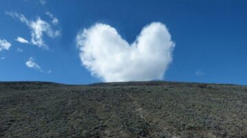 سحابة على شكل قلب تتوسط سماء زرقاء، فوق تلة خضراء. المنظر الطبيعي يعكس جمال الطبيعة والهدوء.