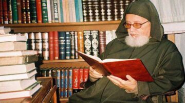 رجل مسن يرتدي عباءة تقليدية ونظارات، يجلس في مكتبة مليئة بالكتب، ويقرأ كتابًا مفتوحًا. الخلفية تحتوي على أرفف مملوءة بالمجلدات، مما يعكس أجواء من الحكمة والمعرفة.