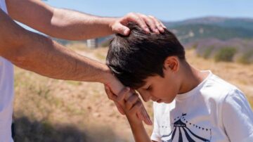 يد تحمل رأس طفل صغير بينما ينحني برأسه إلى الأسفل في منظر طبيعي هادئ. تعكس الصورة مشاعر الحماية والراحة في بيئة طبيعية جميلة، مع تلال خضراء في الخلفية.