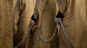 أشخاص يرتدون ألبسة بسيطة من الخيش، مقيدون بسلاسل، تظهر أيديهم من بين الأقمشة، مما يعكس شعورًا بالقبض والاحتجاز.