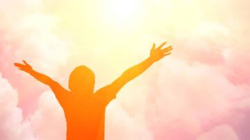 شخص يقف مع ذراعيه ممدودتين نحو السماء، محاطًا بسحب ملونة بألوان دافئة، مما يعكس شعورًا بالحرية والأمل تحت أشعة الشمس.