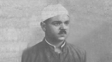 صورة تاريخية لرجل يرتدي قبعة بيضاء وزي تقليدي، يظهر بملامح جادة ونظرة مركزة. الخلفية غير واضحة، مما يبرز ملامح وجهه.