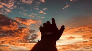 يد مرفوعة نحو سماء ملونة بألوان الغروب، مع سحب تتدرج بين البرتقالي والأزرق. تعكس الصورة شعور الأمل والحرية.