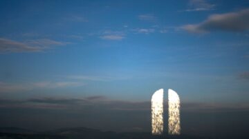 انعكاس ضوء الشمس على نافذة مفتوحة في سماء زرقاء، مما يخلق تأثيراً بصرياً مميزاً. السحب تتوزع بشكل خفيف، مما يضيف عمقاً إلى المشهد الطبيعي.