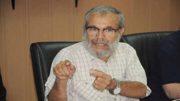 رجل مسن ذو لحية وشعر رمادي يرتدي نظارات وقميص مقلم، يتحدث بحماس أثناء جلوسه في مكتب. تظهر خلفه جدران خشبية ومقاعد جلدية، مما يضفي طابعًا رسميًا على الاجتماع.