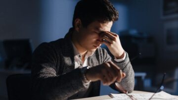 رجل يجلس على مكتب في بيئة مكتبية منخفضة الإضاءة، يمسك نظارته بيده ويضع الأخرى على جبهته، يبدو عليه الإحباط أو التوتر. خلفه جهاز كمبيوتر ومكتب يحتوي على أوراق.