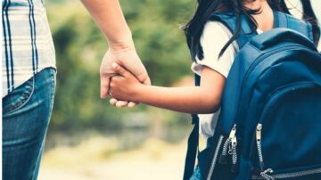 طفلة صغيرة تحمل حقيبة مدرسية تمسك بيد والدها في مشهد يعكس اللحظات العاطفية بين الأهل والأبناء، مع خلفية طبيعية تضفي شعورًا بالأمان والدعم.