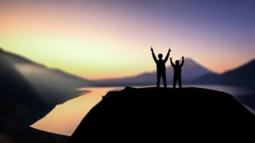 شخصان يقفان على قمة جبل مع رفع أيديهما في الهواء، خلفهما منظر غروب الشمس الجميل على بحيرة وجبال.