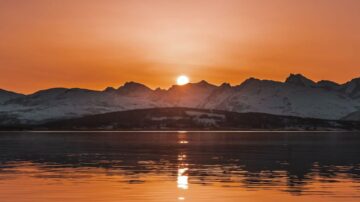 Sunset over snow-capped mountains, reflecting on calm water in a serene landscape. Warm orange and gold hues fill the sky, creating a tranquil atmosphere.