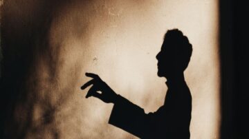 ظل شخص يتحدث بيد مرفوعة، مع خلفية داكنة تضيف جوًا غامضًا. تعكس الصورة الإبداع والتعبير الفني من خلال اللعب بالضوء والظل.