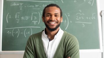 معلم رياضيات مبتسم يقف أمام سبورة خضراء مليئة بالمعادلات الرياضية. يرتدي سترة خضراء وقميص أبيض، ويظهر تعبيره الثقة والحماس للتعليم.