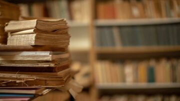 كتب قديمة مكدسة في طاولة، مع خلفية تضم رفوف مليئة بالكتب ذات الأغلفة المتنوعة، تعكس أجواء المكتبات التقليدية.