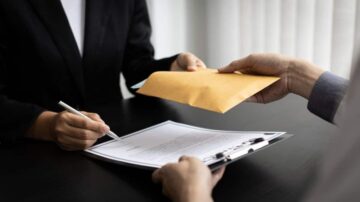 امرأة ترتدي بدلة تسجل معلومات على مستند، بينما يقوم شخص آخر بتسليم ظرف بني يحتوي على مستندات. المشهد يعكس لحظة تبادل الوثائق الرسمية في بيئة مكتبية.