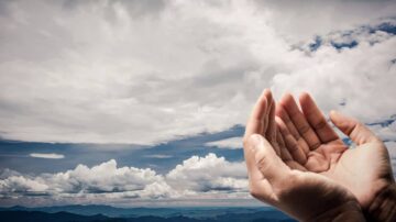 يدين مفتوحتان نحو السماء المليئة بالغيوم، مع خلفية جبال بعيدة. تعبر الصورة عن الأمل والرحمة والتواصل الروحي.