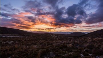 غروب الشمس فوق تلال مغطاة بالنباتات، مع سماء ملبدة بالغيوم تتلون بألوان دافئة. منظر طبيعي خلاب يعكس جمال الطبيعة في الصحراء.