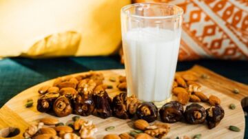 كوب من الحليب في وسط صحن خشبي محاط بالتمر والمكسرات مثل اللوز والجوز، مع خلفية ملونة تضفي جوًا دافئًا.