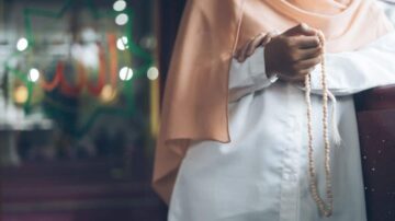 امرأة ترتدي حجابًا برتقاليًا، تمسك بمسبحة في يدها، خلفها خلفية تضئ بكلمات دينية تشير إلى الإيمان. تعبر الصورة عن روحانية وتأمل.