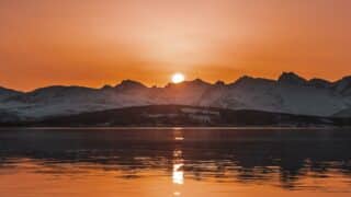 Sunset over snowy mountains, reflecting on a calm lake, in warm orange colors.