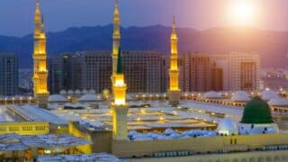 Large mosque with illuminated minarets and green dome at dusk, mountains in background.