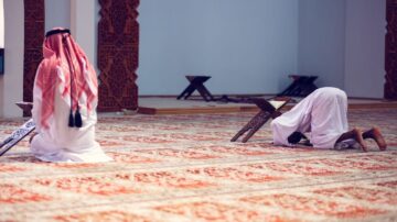 صورة لمصلين يرتديان الملابس التقليدية في مسجد، حيث يجلس أحدهم على ركبتيه والآخر يسجد. تظهر خلفية المسجد تفاصيل زخرفية وأرضية مفروشة بالسجاد.