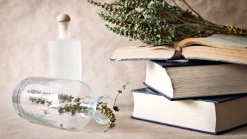 كتابان مفتوحان وكتاب مغلق موضوعة فوق طاولة، مع زجاجة شفافة تحتوي على أعشاب جافة ووعاء زجاجي مائل. خلفية بنية تضفي جوًا دافئًا.