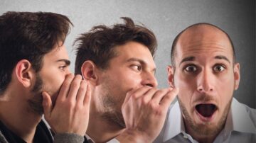 ثلاثة رجال يظهرون في الصورة. الرجل الأول يستمع بتركيز، بينما الثاني يهمس في أذنه. الرجل الثالث يبدو متفاجئًا أو مصدومًا. الخلفية رمادية، مما يبرز تعابير الوجه. تعكس الصورة تفاعلًا اجتماعيًا قويًا.