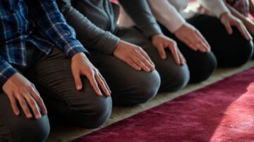 مجموعة من الأشخاص يجلسون على الأرض في وضعية الصلاة، مع التركيز على أيديهم المتكئة على ركبهم، والخلفية تُظهر سجادة صلاة حمراء.